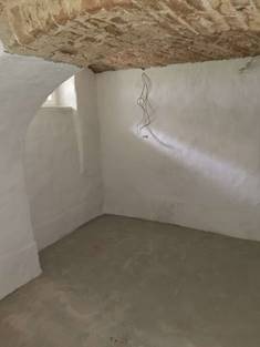 Wnętrze piwnicy z surowym, ceglanym sklepieniem łukowym, świeżo wylaną betonową posadzką i otynkowanymi ścianami, widoczne przewody elektryczne zwisające ze ściany, małe okno w łuku.