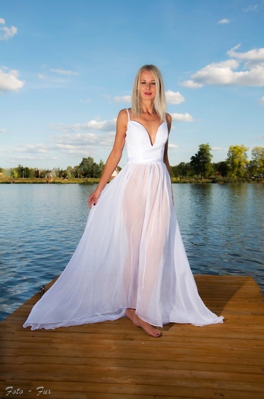 Kobieta w zwiewnej białej sukni na drewnianym pomoście nad jeziorem, błękitne niebo w tle.