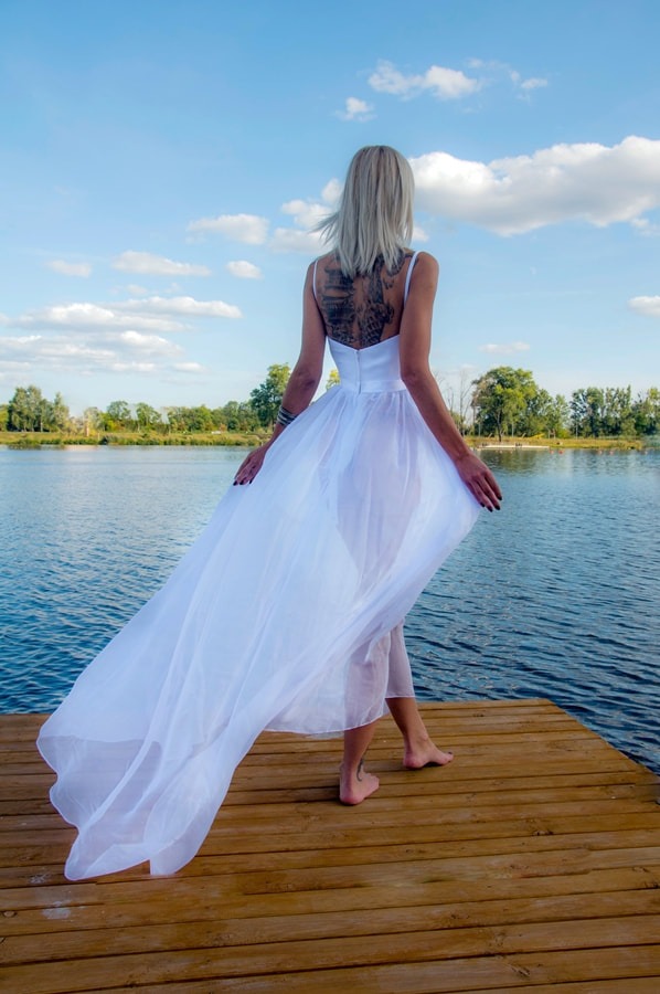 Kobieta w zwiewnej białej sukience, stojąca tyłem na drewnianym pomoście nad wodą, z tatuażem na plecach, patrząca w dal na jezioro i drzewa.