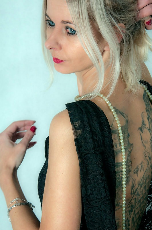 Portret kobiety z blond włosami, perłami i tatuażem na plecach, ubrana w czarną koronkową sukienkę, spoglądająca w lewo.
