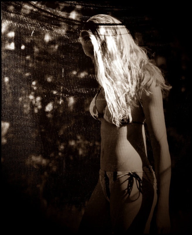 Kobieta w bikini, częściowo zasłonięta siatką, w sepia, z widocznym zarysem sylwetki i oświetleniem podkreślającym kształty.