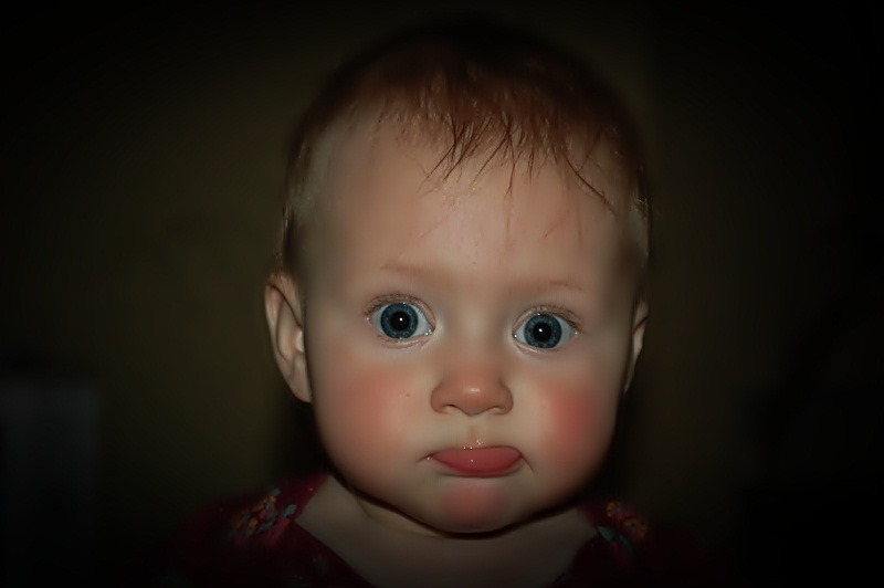 Portret niemowlęcia z wyrazistymi niebieskimi oczami i lekko wysuniętym językiem na ciemnym tle.