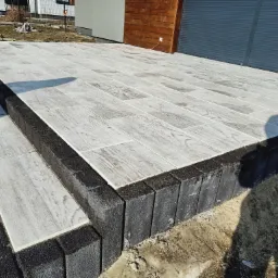 Jasnoszara, wyłożona płytami tarasowymi powierzchnia z obramowaniem z ciemnych, prostokątnych kostek, prowadząca do budynku z elementami drewnianymi i szarą roletą.