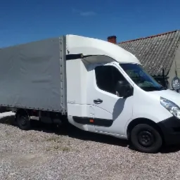 Biały bus dostawczy z szarą plandeką na żwirowym podłożu, widok z boku, w tle budynek z anteną satelitarną.