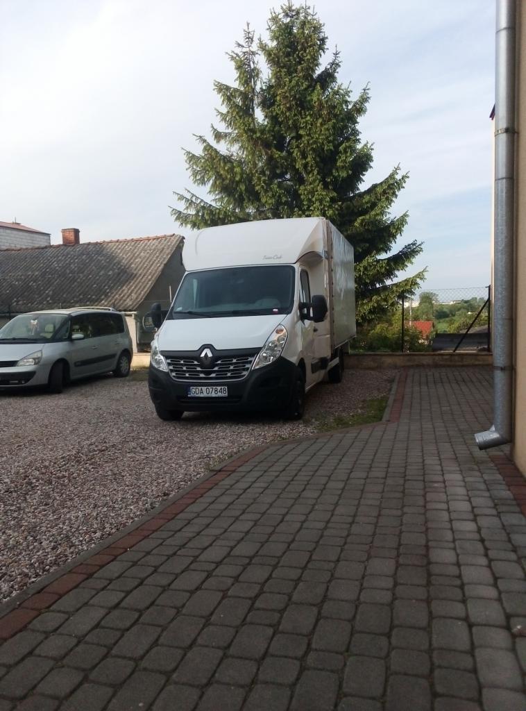 Biały samochód dostawczy Renault Master z zabudową kontenerową zaparkowany na posesji z brukowaną ścieżką i żwirowym podłożem, na tle zielonego świerku.