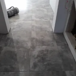 Ułożone szare płytki podłogowe z odcieniem betonu, widoczny boombox w rogu pomieszczenia i fragment futryny drzwiowej.