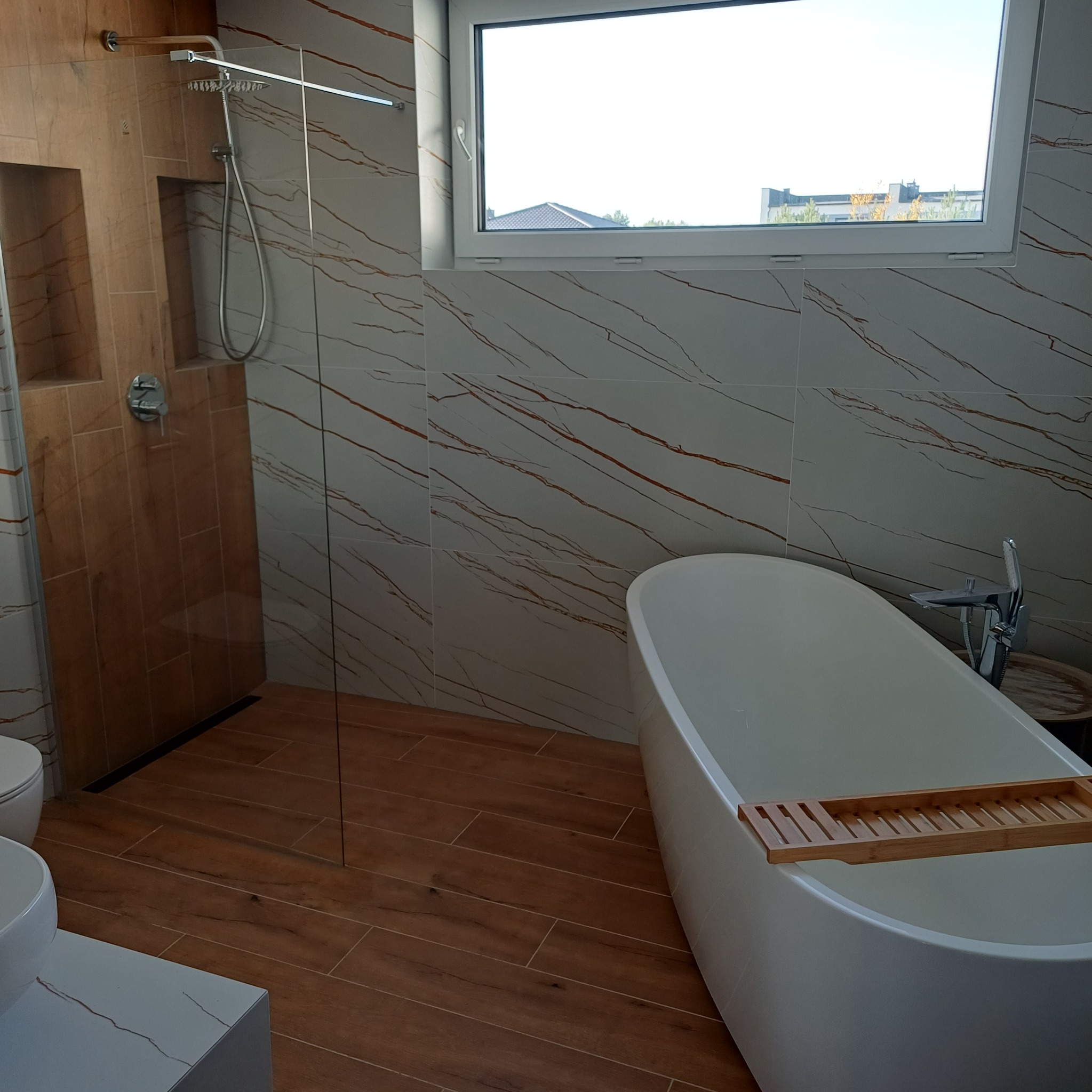 Nowoczesna łazienka z wanną wolnostojącą, kabiną prysznicową typu walk-in i oknem. Płytki imitujące marmur z brązowymi żyłami na ścianach i drewnopodobne na podłodze.