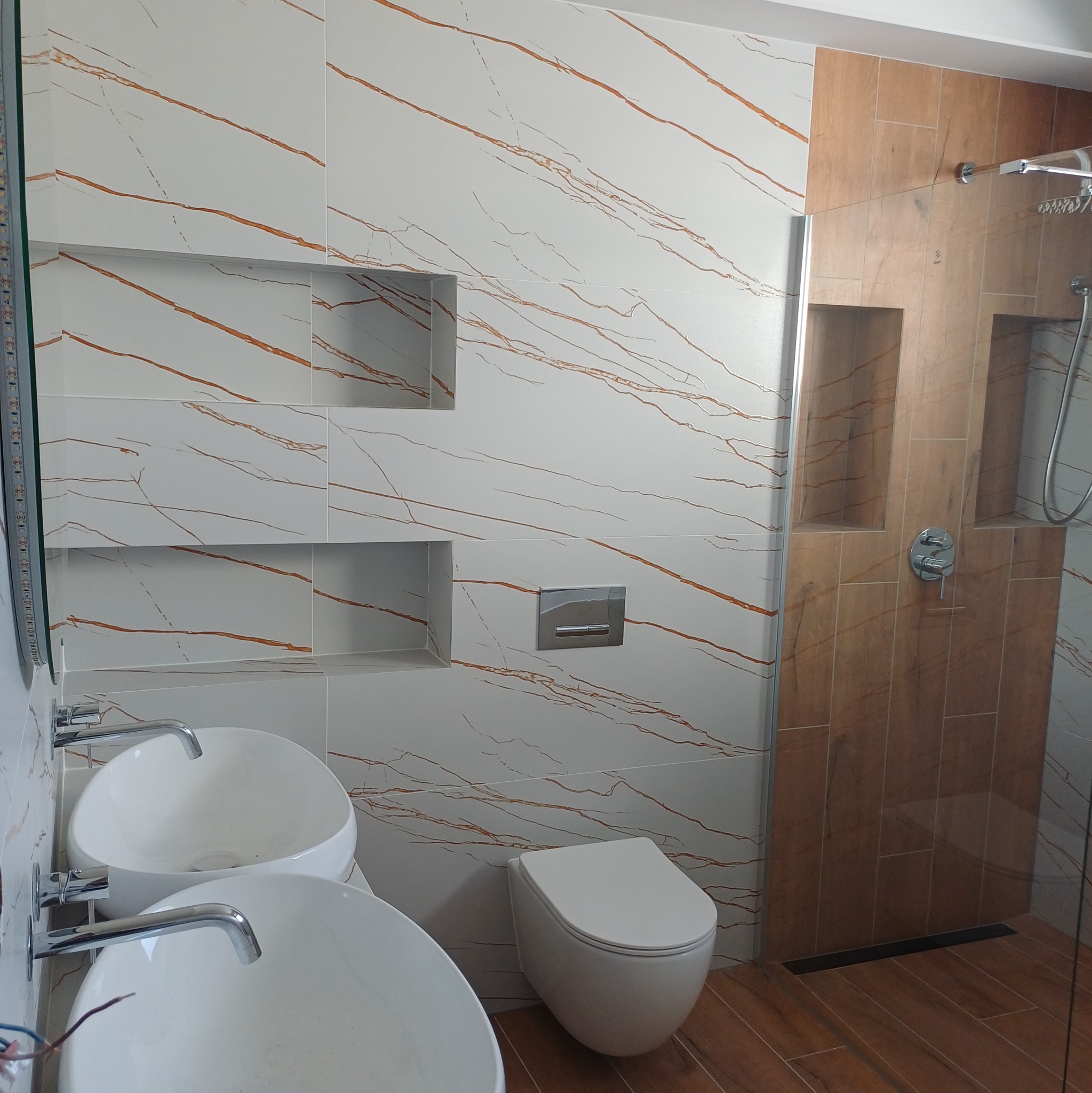 Nowoczesna łazienka z białymi umywalkami nablatowymi, toaletą i kabiną prysznicową. Ściany wyłożone płytkami imitującymi marmur z brązowymi żyłami i drewnopodobnymi.