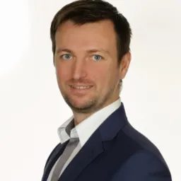 Portret uśmiechniętego mężczyzny w granatowym garniturze, białej koszuli i szarym krawacie na jasnym tle.