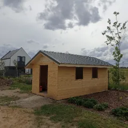 Surowy drewniany domek narzędziowy z blachy falistej na dachu, widoczny z zewnątrz na tle nowego osiedla i pochmurnego nieba.