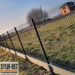 Długi, czarny, druciany system ogradzający na betonowej podmurówce, rozciągający się wzdłuż działki z budynkiem w tle i logo firmy w lewym dolnym rogu.