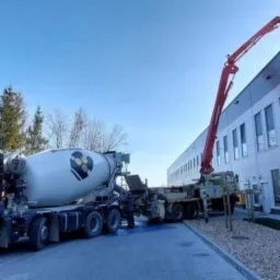 Ciężarówka z betonomieszarką oraz pompa do betonu podczas wylewania betonu na budowie budynku biurowego w słoneczny dzień.