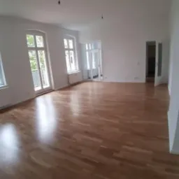 Jasne, przestronne wnętrze z parkietem ułożonym w jodełkę, widoczne okna balkonowe i drzwi do kolejnych pomieszczeń, ściany pomalowane na biało, brak mebli.