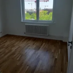 Pusty pokój z parkietem w jodełkę, białe ściany, grzejnik pod oknem z widokiem na budowę z dźwigami.