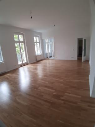 Jasne, przestronne wnętrze z parkietem ułożonym w jodełkę, widoczne okna balkonowe i drzwi do kolejnych pomieszczeń, ściany pomalowane na biało, brak mebli.