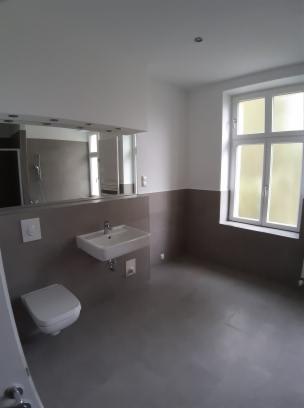 Wykończona łazienka z szarą podłogą, białą umywalką, toaletą i lustrem z oświetleniem, widok z perspektywy drzwi.