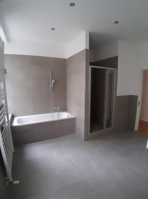 Wykończona łazienka z szarymi płytkami, białą wanną i grzejnikiem drabinkowym, widoczna kabina prysznicowa z przezroczystymi drzwiami.