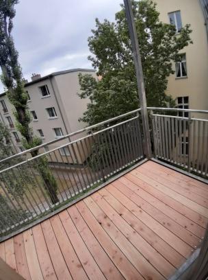 Nowoczesny balkon z drewnianą podłogą i metalową balustradą, widok z góry na tle budynków mieszkalnych i drzew.