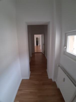Wnętrze korytarza z drewnianą podłogą i białymi ścianami, widok w głąb z oknem po prawej stronie i grzejnikiem pod nim.