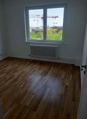 Pusty pokój z parkietem w jodełkę, białe ściany, grzejnik pod oknem z widokiem na budowę z dźwigami.