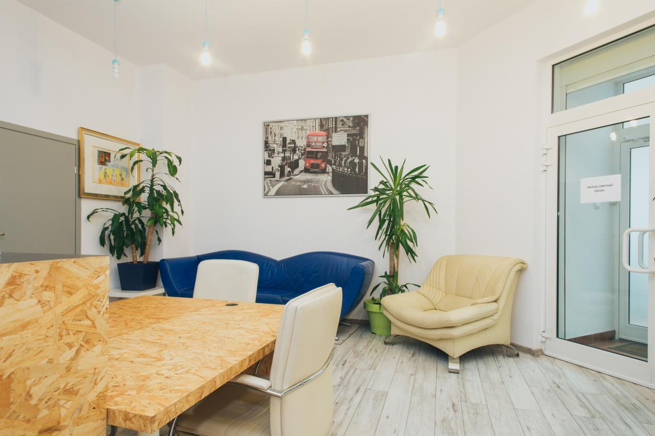 Jasne, przestronne wnętrze biura z drewnianym stołem z płyty OSB, niebieską sofą, kremowym fotelem, roślinami w doniczkach i obrazem z czerwonym autobusem w Londynie na ścianie.