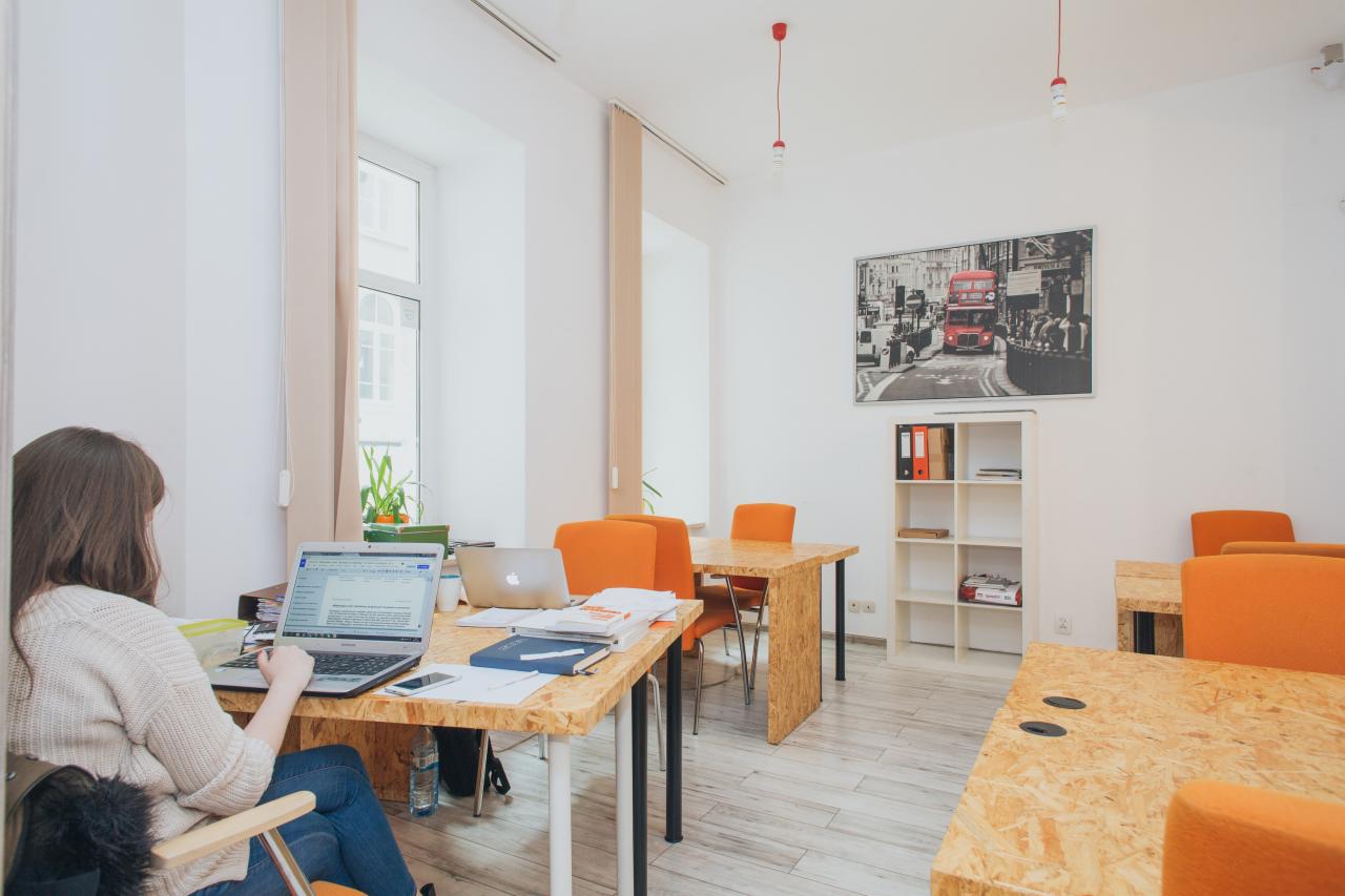 Kobieta pracuje na laptopie w jasnym biurze wirtualnym z pomarańczowymi krzesłami i stolikami z płyty OSB, na ścianie duża grafika z czerwonym autobusem.