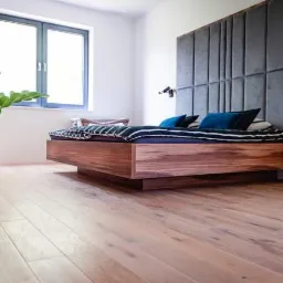 Nowoczesna sypialnia z drewnianym łóżkiem i wezgłowiem z szarego, tapicerowanego panelu. Minimalistyczne oświetlenie i drewniana podłoga.