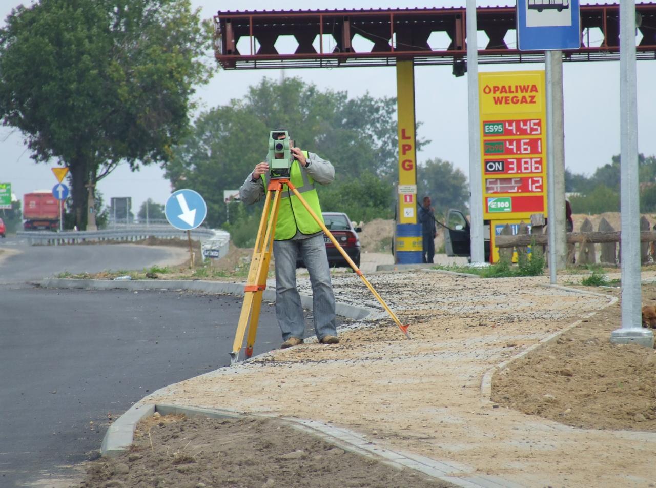 Pracownik w kamizelce odblaskowej obsługuje teodolit na trójnogu przy budowie drogi w pobliżu stacji benzynowej.