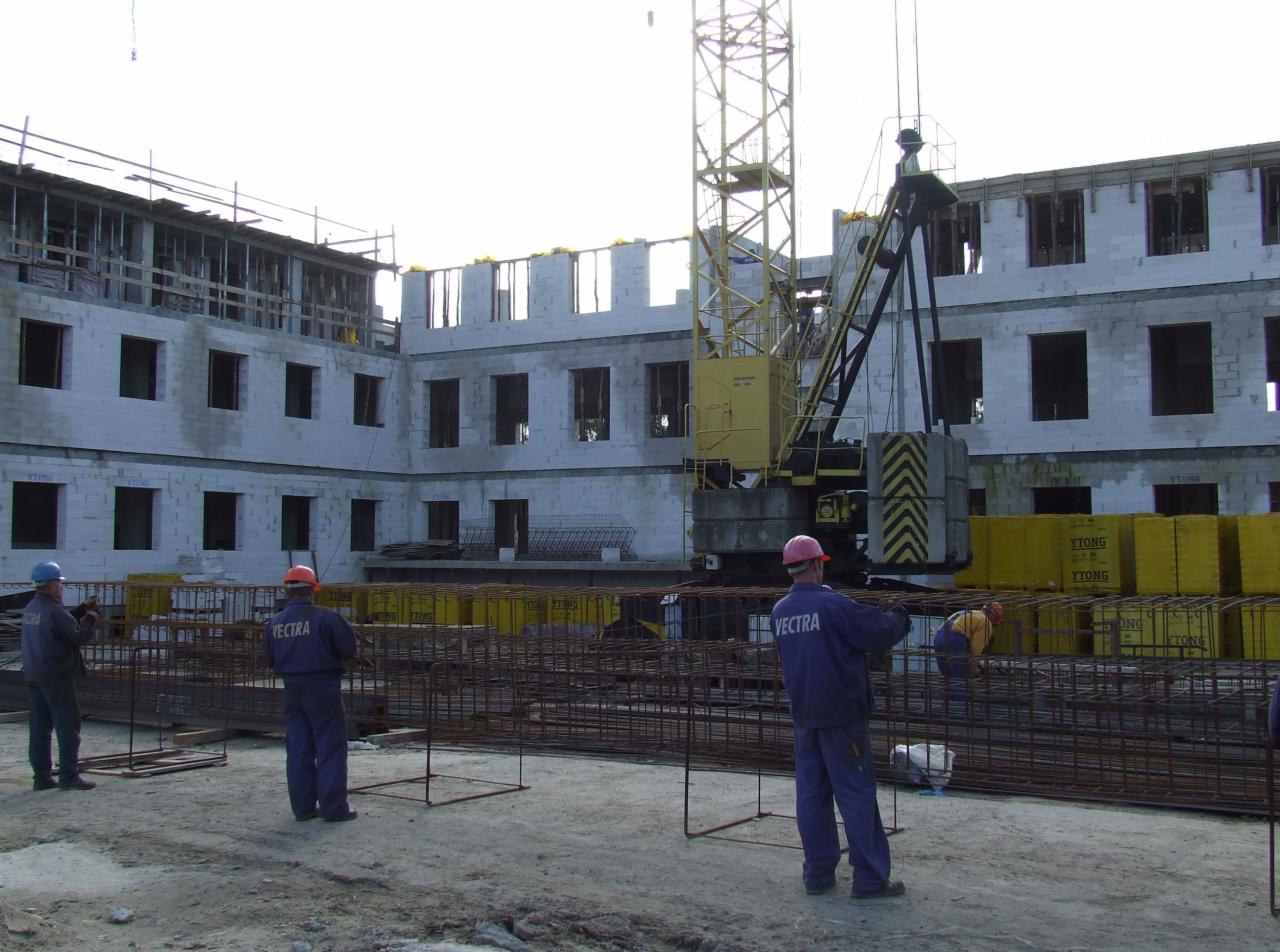 Budowa wielopiętrowego budynku z białego bloczka YTONG, widoczny żółty dźwig budowlany oraz pracownicy w niebieskich kombinezonach z napisem VECTRA, przygotowujący zbrojenie.