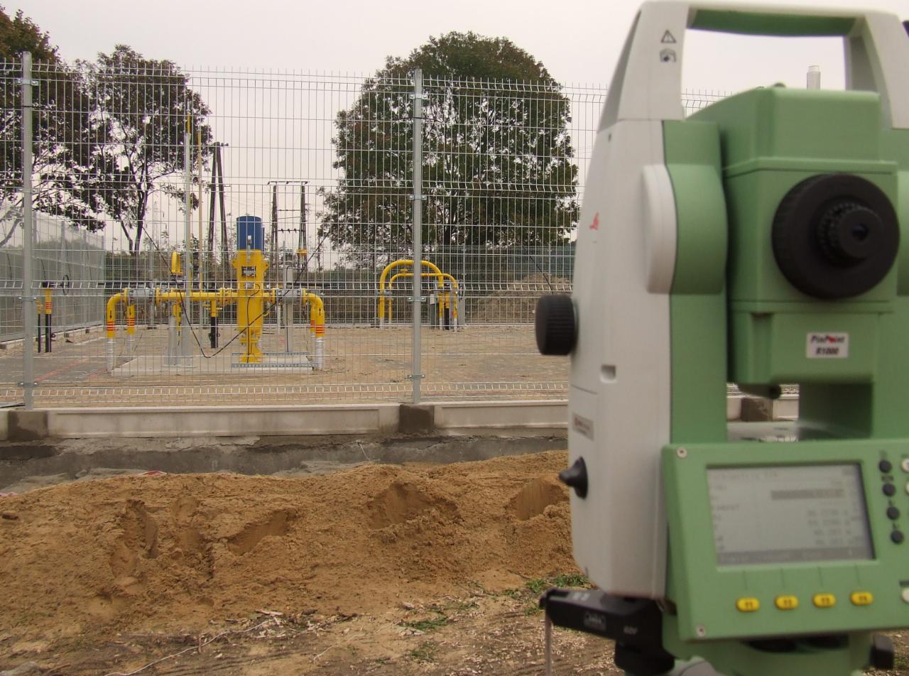 Profesjonalny niwelator optyczny Leica na statywie, z widoczną żółtą instalacją gazową w tle, oddzieloną metalową siatką.