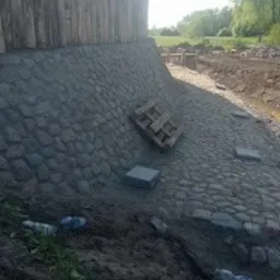 Skarpa wyłożona szarą kostką brukową przy drewnianej konstrukcji. Na kostce leży drewniana paleta i kilka płyt chodnikowych. U dołu widoczne śmieci: butelki plastikowe.