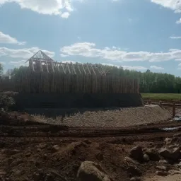 Budowa drewnianej palisady z konstrukcją dachu w trakcie realizacji, widoczne przygotowane podłoże i otaczający teren.