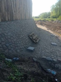 Skarpa wyłożona szarą kostką brukową przy drewnianej konstrukcji. Na kostce leży drewniana paleta i kilka płyt chodnikowych. U dołu widoczne śmieci: butelki plastikowe.