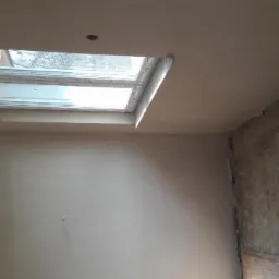 Wnętrze pokoju po nałożeniu gładzi gipsowej, widoczne okno dachowe i fragment ściany z folią ochronną.