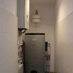 Kompaktowa kotłownia w minimalistycznym stylu: białe płytki, szary zasobnik ciepłej wody z czarnym panelem rewizyjnym, biały wiszący piec gazowy, widoczne rury i zawory.