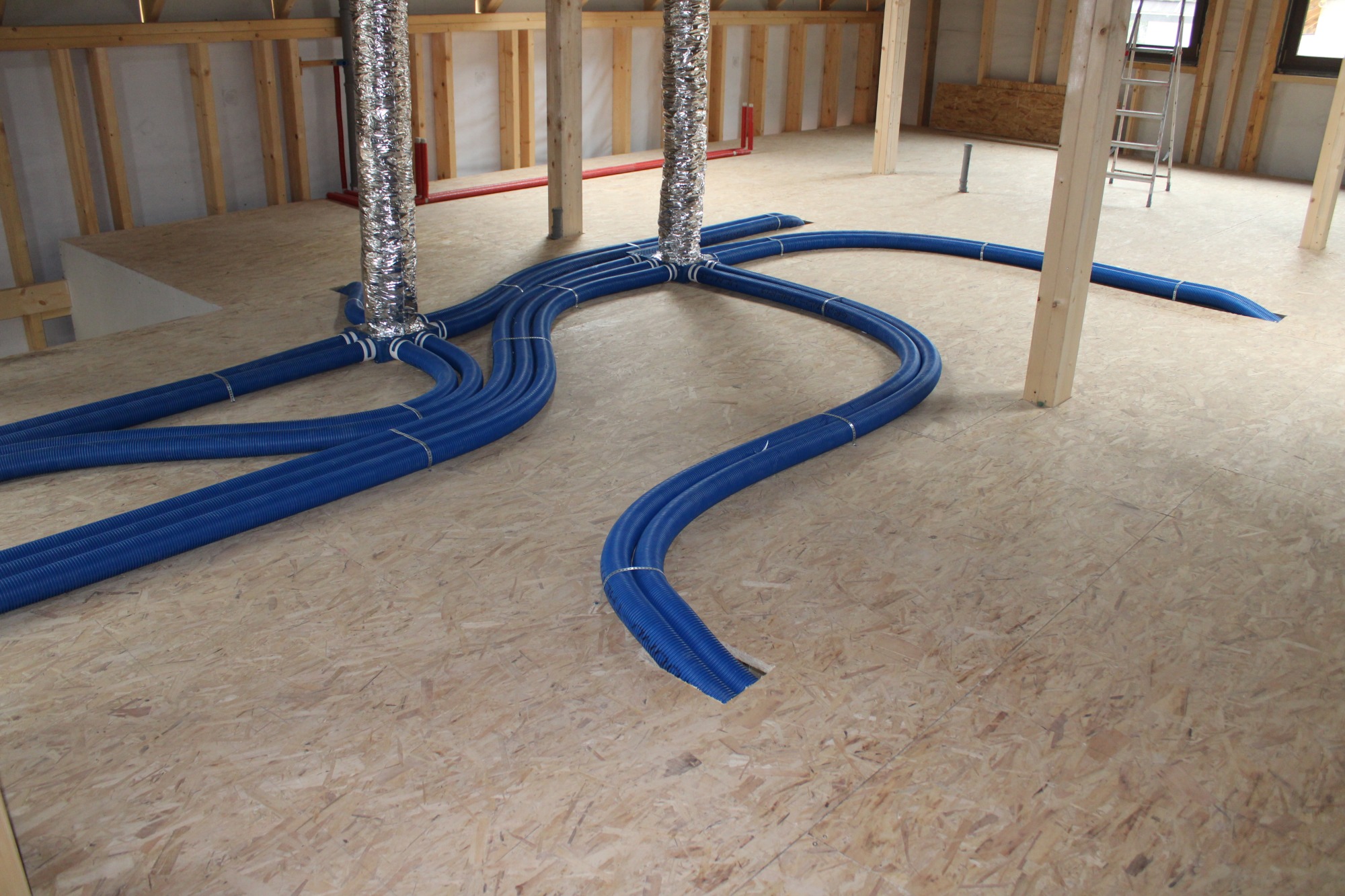 Niebieskie, karbowane rury wentylacyjne rozchodzą się promieniście od pionowych, aluminiowych przewodów na podłodze z płyt OSB w surowym wnętrzu budynku w trakcie budowy.