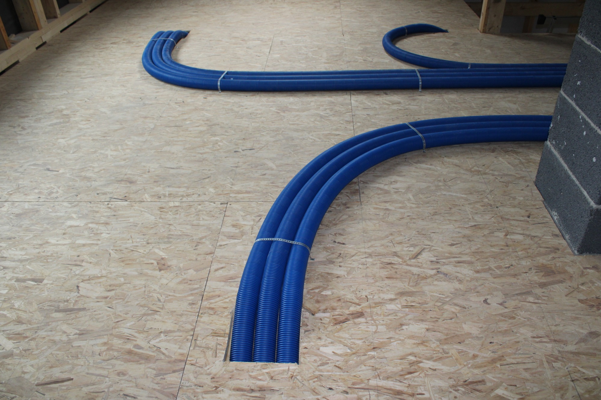 Niebieskie, karbowane rury ogrzewania podłogowego prowadzone w wycięciach płyty OSB, spięte metalowymi opaskami, widok z góry, fragment ściany z bloczków betonowych w tle.