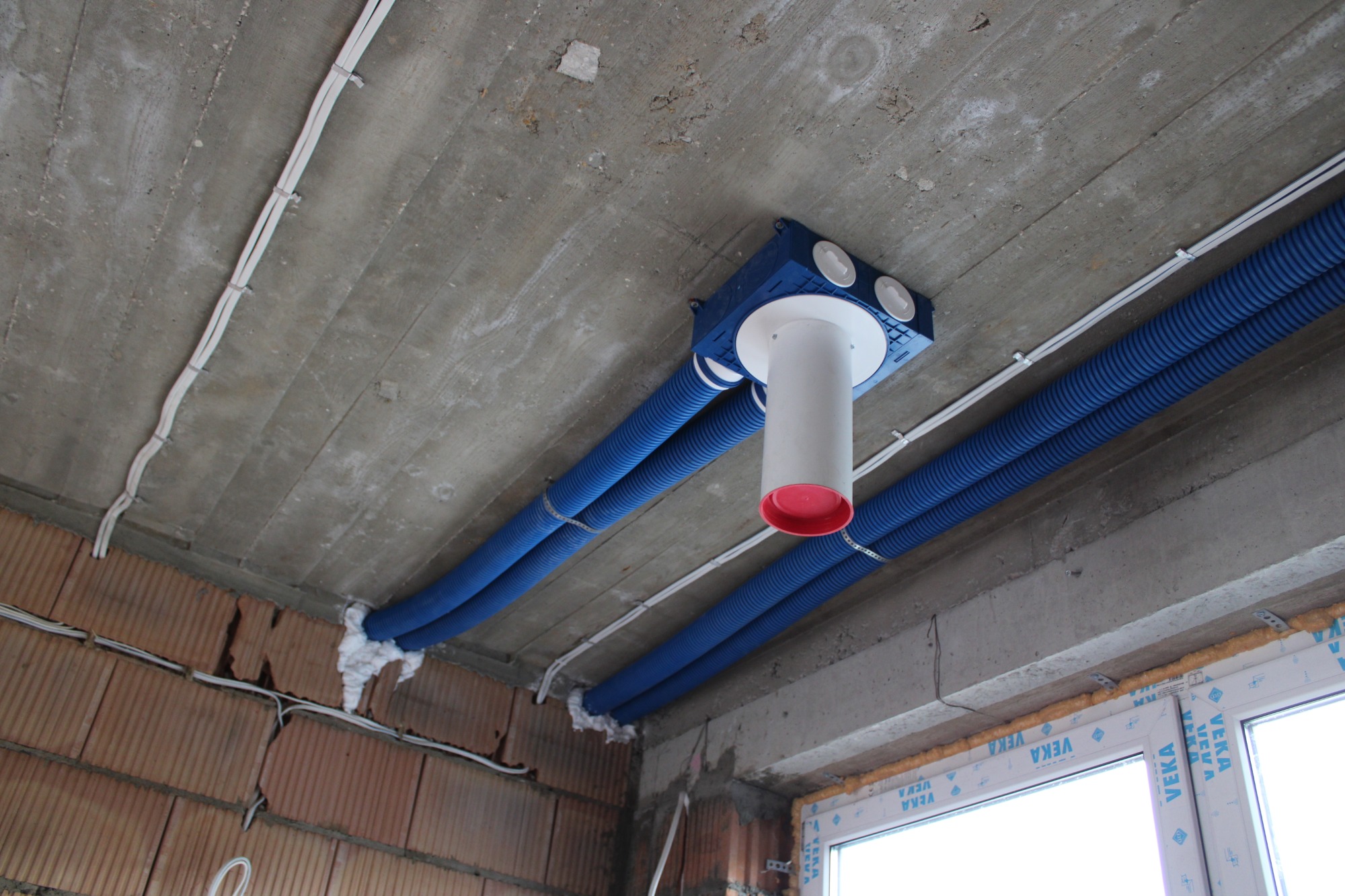 Niebieskie rury wentylacyjne przymocowane do surowego betonowego sufitu, z białym pionowym kanałem zakończonym czerwonym pierścieniem, wychodzącym z niebieskiej skrzynki rozdzielczej.