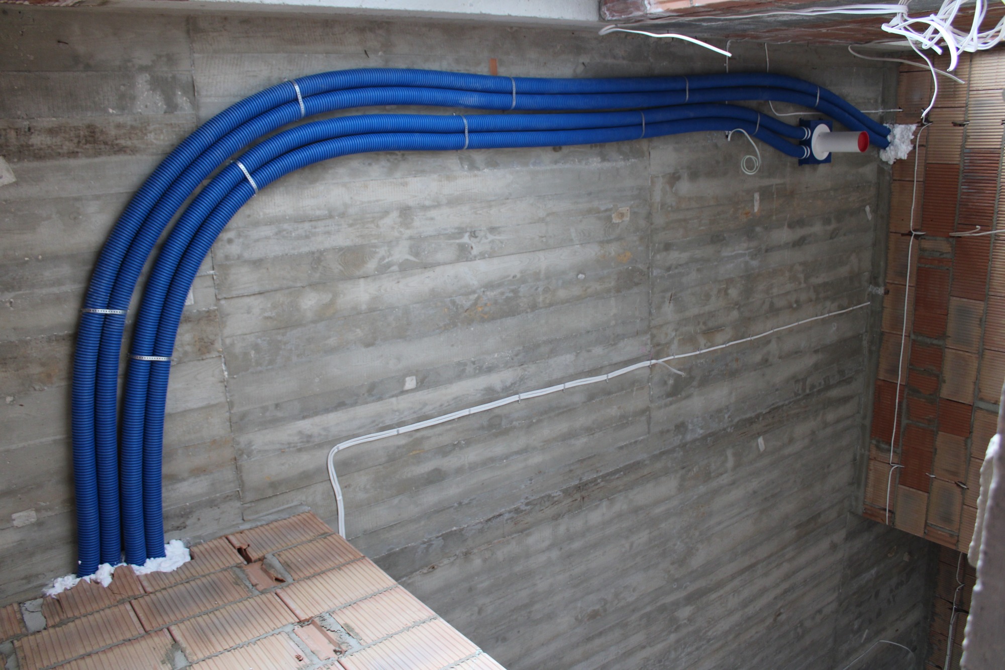 Niebieskie, karbowane rury wentylacyjne przymocowane do surowej betonowej ściany w Grabicy, z widocznymi przewodami elektrycznymi i fragmentem muru z cegieł.