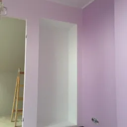 Świeżo pomalowane na liliowo ściany z wnęką pomalowaną na biało, widoczna drabina w tle, listwy przypodłogowe w kolorze ciemnego brązu.