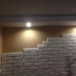 Częściowo wykończona ściana z dekoracyjnej cegły w odcieniach bieli i szarości, oświetlona punktowo, z widocznym fragmentem drabiny i beżową ścianą w tle.