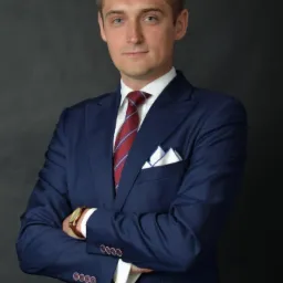 Portret mężczyzny w granatowym garniturze, z założonymi rękami. Biała koszula, bordowy krawat w paski i poszetka w kieszonce dodają elegancji. Ciemne tło.