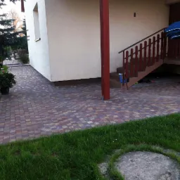 Chodnik z kostki brukowej w odcieniach brązu i czerwieni, otoczony zielonym trawnikiem, prowadzący do schodów wejściowych domu z drewnianą kolumną.