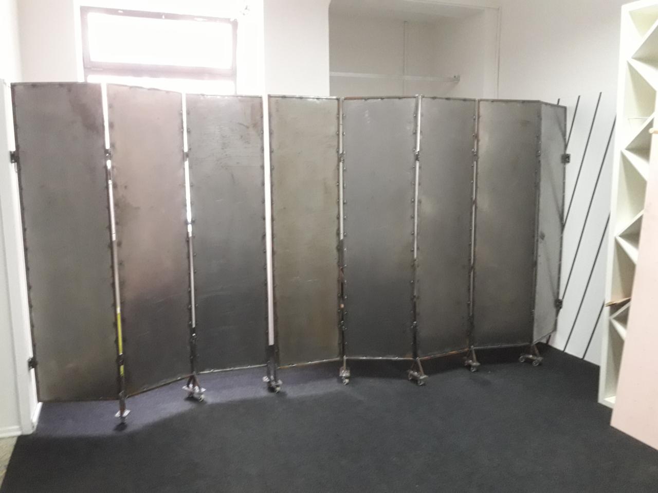 Składany, metalowy parawan z siedmioma segmentami na kółkach, ustawiony w jasnym pomieszczeniu z oknem.