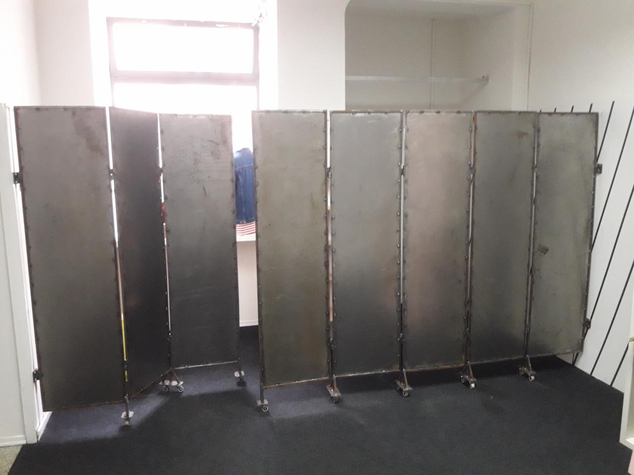Metalowy parawan pokojowy z siedmioma segmentami, na kółkach, spawany, w surowym wykończeniu, ustawiony na czarnej wykładzinie.