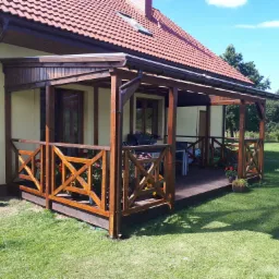 Zadaszony taras z drewna przy domu jednorodzinnym, z drewnianą balustradą zdobioną kwiatami w donicach, grill i stół z krzesłami na tle zielonego trawnika.