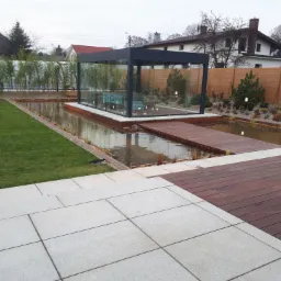 Nowoczesna, przeszklona konstrukcja z czarną ramą, umieszczona na platformie otoczonej wodą, z drewnianym pomostem, widok z tarasu z jasnych płyt, w tle zielony trawnik i płot.