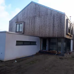 Nowoczesny dom o prostej bryle, z drewnianą elewacją z pionowych desek i dużymi przeszkleniami, w trakcie budowy, z widocznym stosem drewna przed wejściem i ziemistym terenem wokół.