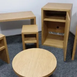 Zestaw mebli z jasnego drewna: stolik kawowy okrągły, szafka z otwartymi półkami i drzwiczkami, mały stolik nocny z półkami, oraz wąski stolik pomocniczy z półkami, ustawione na szarym dywanie.