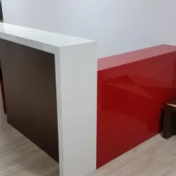 Nowoczesna lada recepcyjna z białym blatem, brązowym panelem z logo w czerwonej obw&oacute;dce i czerwoną, lakierowaną ścianką boczną, uzupełniona stolikiem kawowym w geometryczne wzory.
